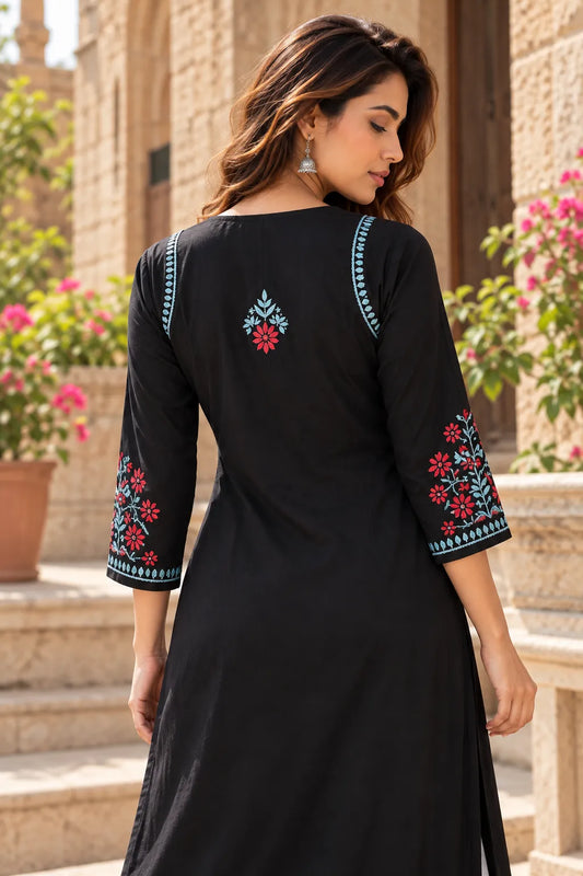 A woman wearing a black three-quarter sleeve kurta with colorful floral embroidery on the sleeves and back, photographed outdoors in front of a stone building.