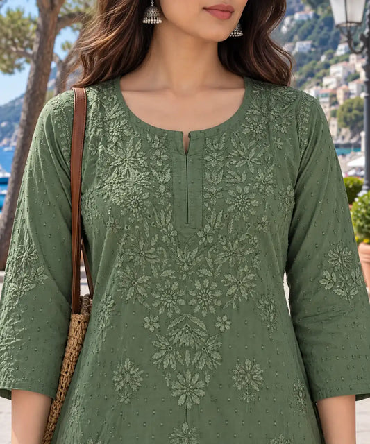 A woman wearing a sage green embroidered kurta with floral lace detailing, photographed outdoors near a coastal town with mountains and water in the background.