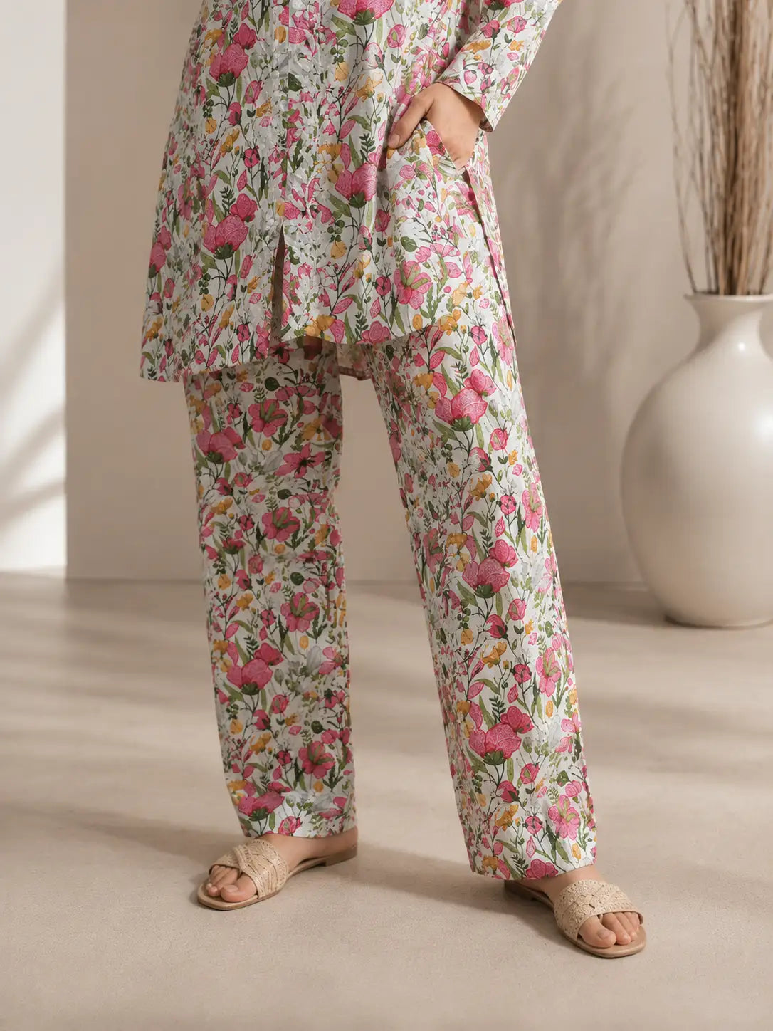 A person wearing a matching floral long-sleeve tunic top and wide-leg pants set in a soft pink, green, and white print, styled with beige slide sandals in a warm neutral interior setting.