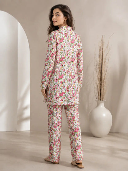 A woman modeling a two-piece floral pajama set, shown from the back. The set features a long-sleeve tunic and matching pants in a light cream base with a pink, red, and green floral pattern.