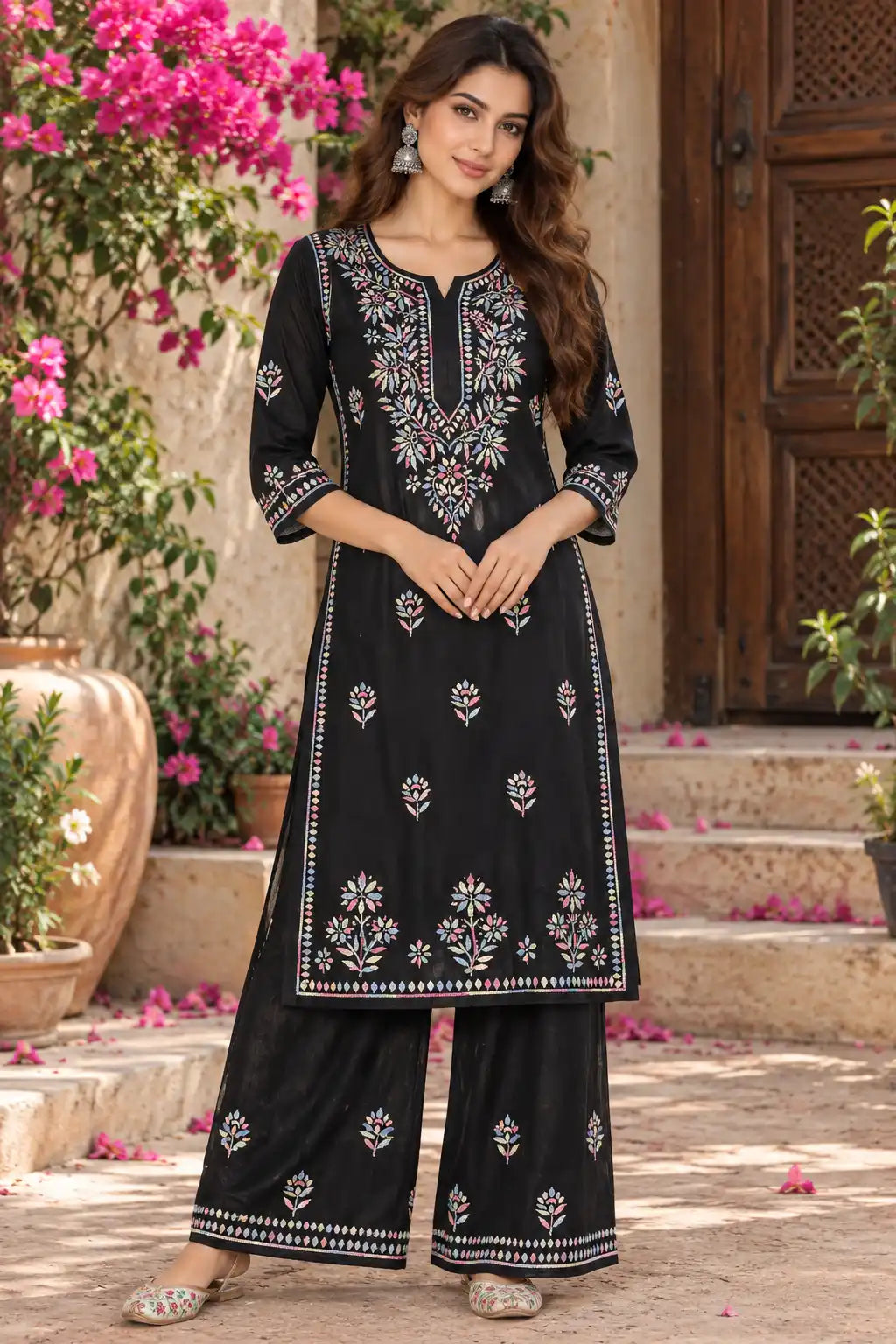 A woman models a black three-piece kurta set with white and pink floral embroidery, standing in front of a rustic stone wall and wooden door with pink flowers in the background.