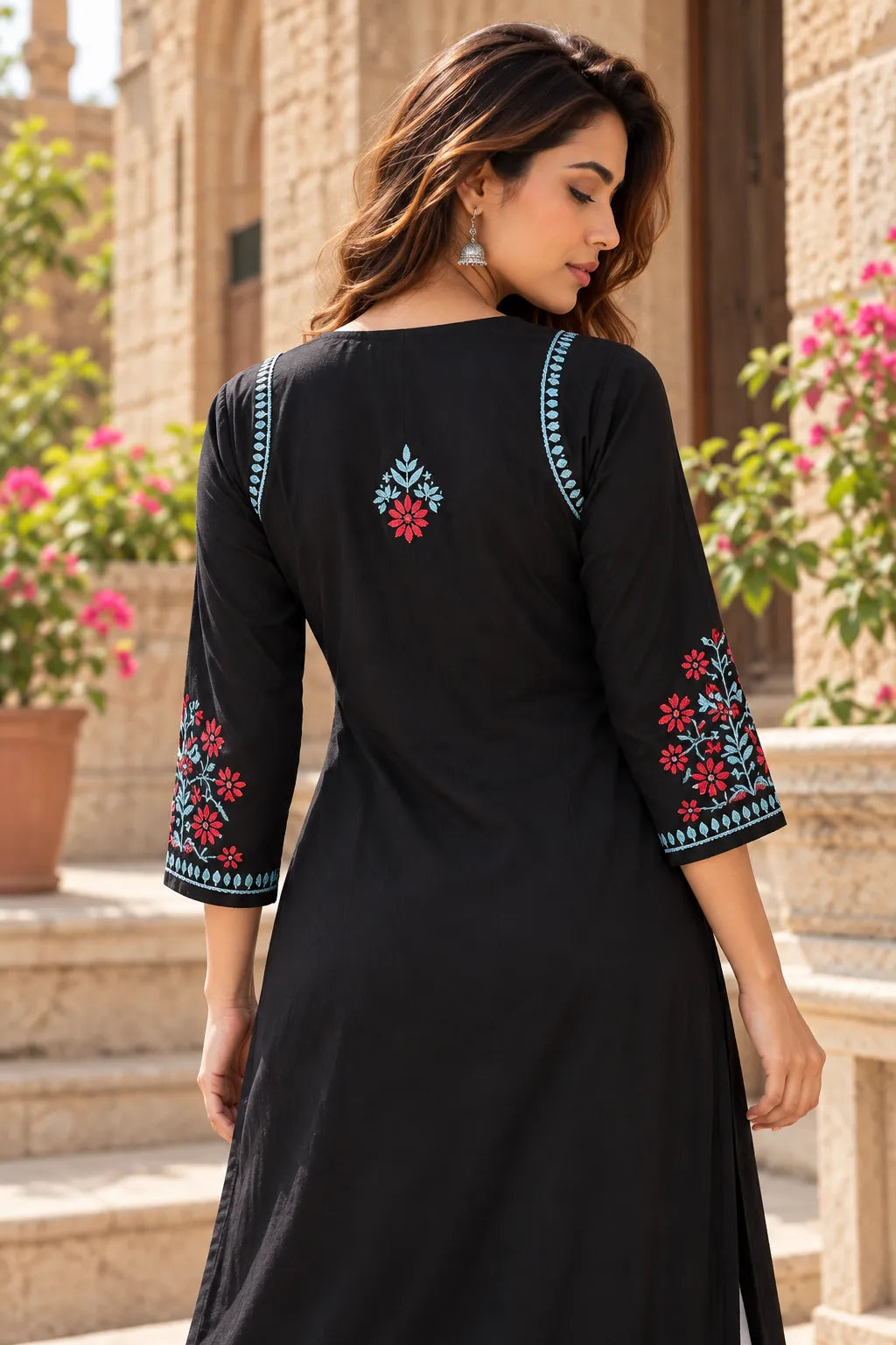 A woman wearing a black three-quarter sleeve kurta with colorful floral embroidery on the sleeves and back, photographed outdoors in front of a stone building.