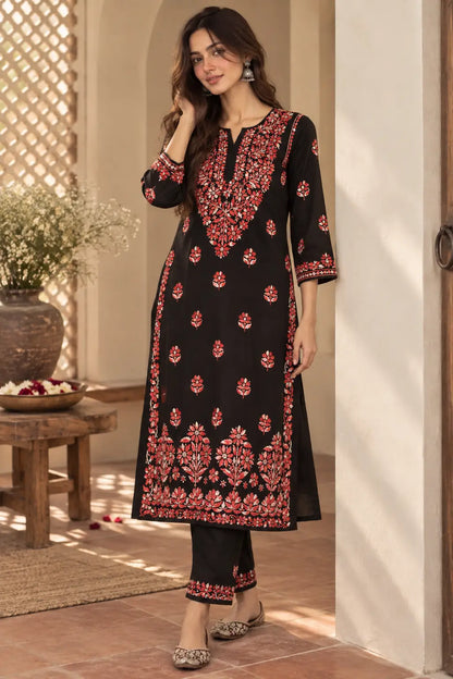 A woman models a black three-piece salwar kameez suit with red and gold floral embroidery, standing in a warm, sunlit traditional courtyard setting.
