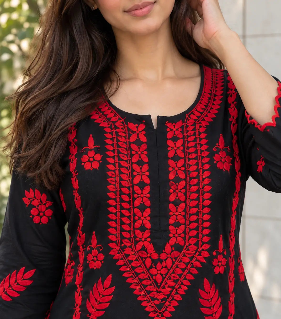 A close-up of a woman wearing a black long-sleeve kurta with vibrant red floral and geometric embroidery, set against a softly blurred outdoor background.