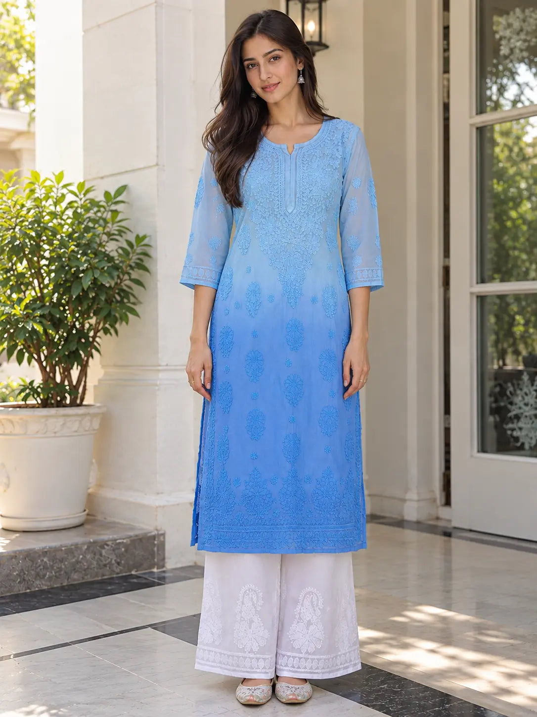 A woman modeling a light blue embroidered kurta with white palazzo pants, standing in front of a white building entrance with a potted plant.
