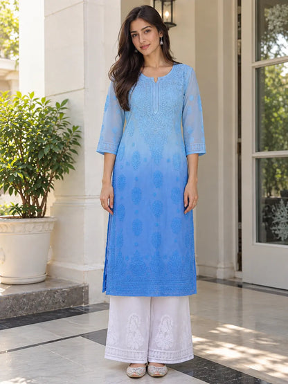 A woman modeling a light blue embroidered kurta with white palazzo pants, standing in front of a white building entrance with a potted plant.