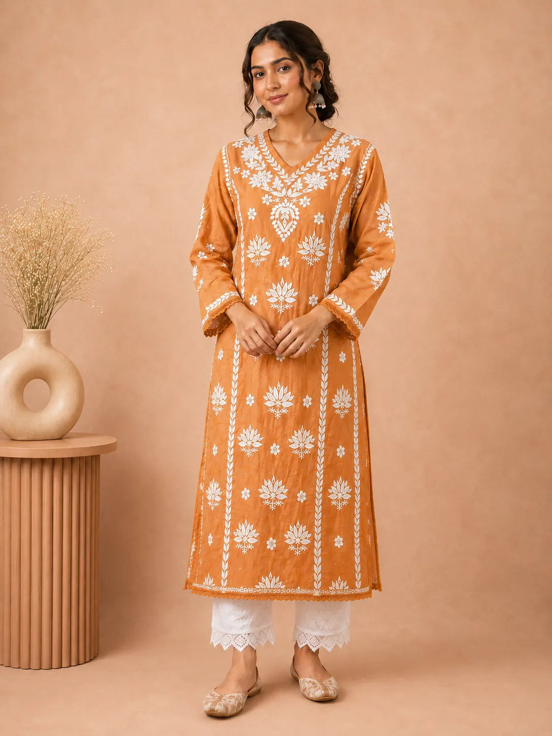 A woman models a mustard orange kurta with white floral embroidery, paired with white pants and beige shoes, set against a warm beige background with a decorative vase on a side table.