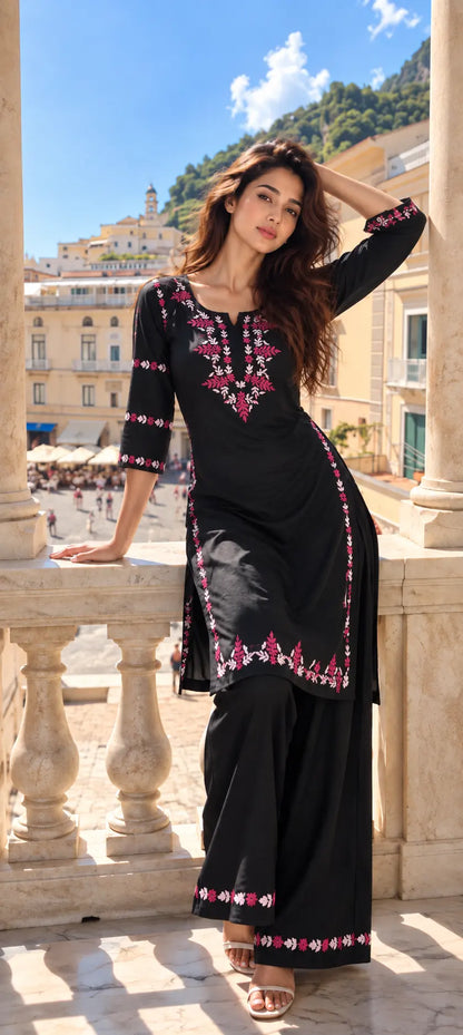 A woman modeling a black three-piece kurta set with pink and white floral embroidery, standing on a stone balcony with a Mediterranean town in the background.