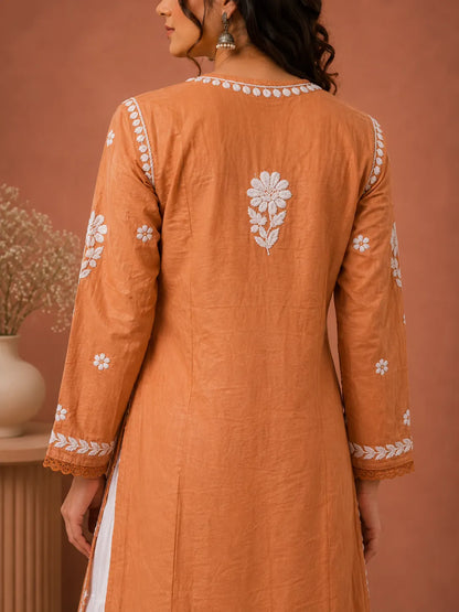 A woman shown from the back wearing a peach-orange long kurta with white floral embroidery on the back, shoulders, and cuffs, set against a warm terracotta background.