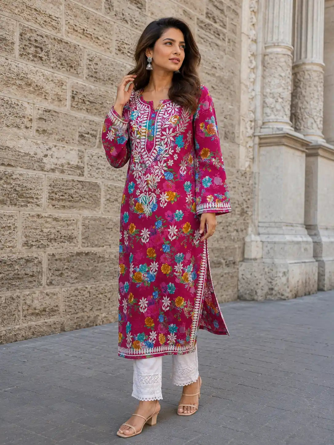 A woman models a vibrant magenta pink embroidered kurta with colorful floral patterns, paired with white pants and beige sandals, standing against a stone wall.