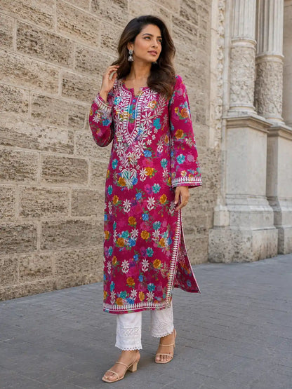 A woman models a vibrant magenta pink embroidered kurta with colorful floral patterns, paired with white pants and beige sandals, standing against a stone wall.