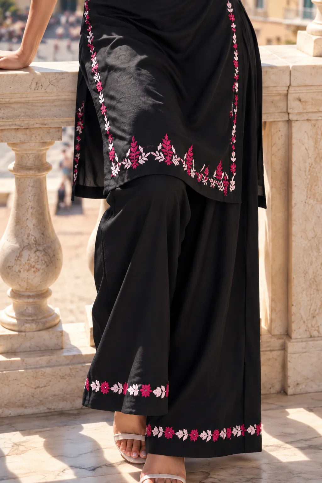 A close-up of a person wearing a black traditional outfit with pink and white floral embroidery along the edges, standing outdoors near a stone balustrade.