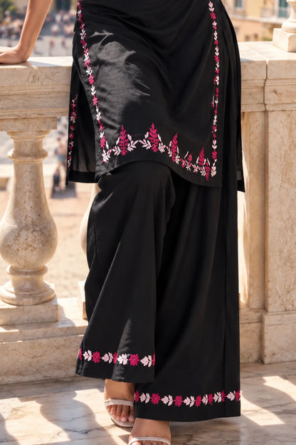 A close-up of a person wearing a black traditional outfit with pink and white floral embroidery along the edges, standing outdoors near a stone balustrade.