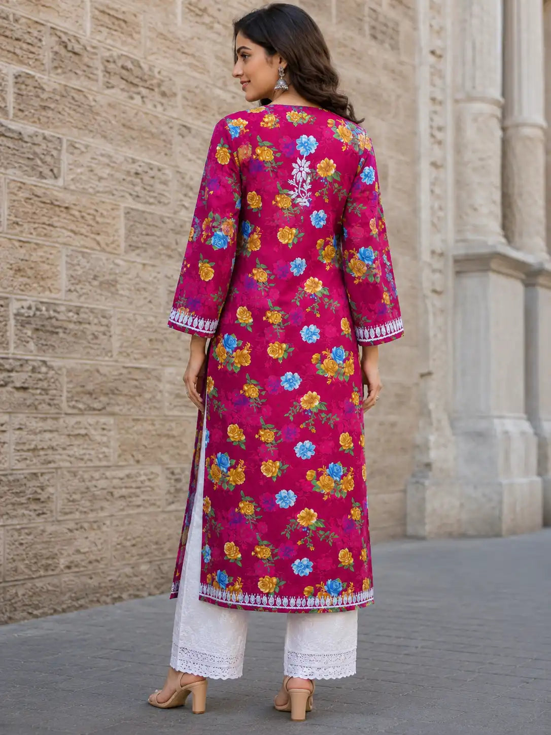 A woman stands outdoors against a stone wall wearing a bright pink embroidered kurta with multicolored floral patterns, paired with white pants and beige heels.