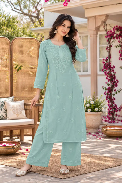 A woman models a mint green embroidered kurta and trouser set in an outdoor patio setting with a wicker screen, cushions, and potted plants.