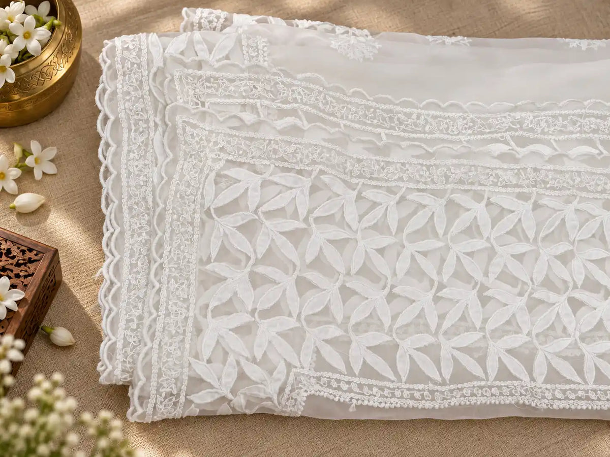 White embroidered lace fabric with a geometric floral pattern and scalloped borders, laid flat on a beige textured surface with decorative flowers and a small wooden box.