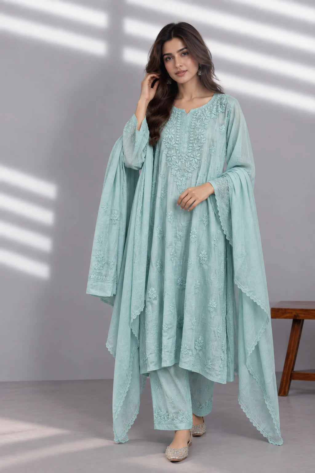 A woman models a light mint green embroidered traditional outfit with a matching draped outer layer, set against a gray wall with soft window shadow patterns.
