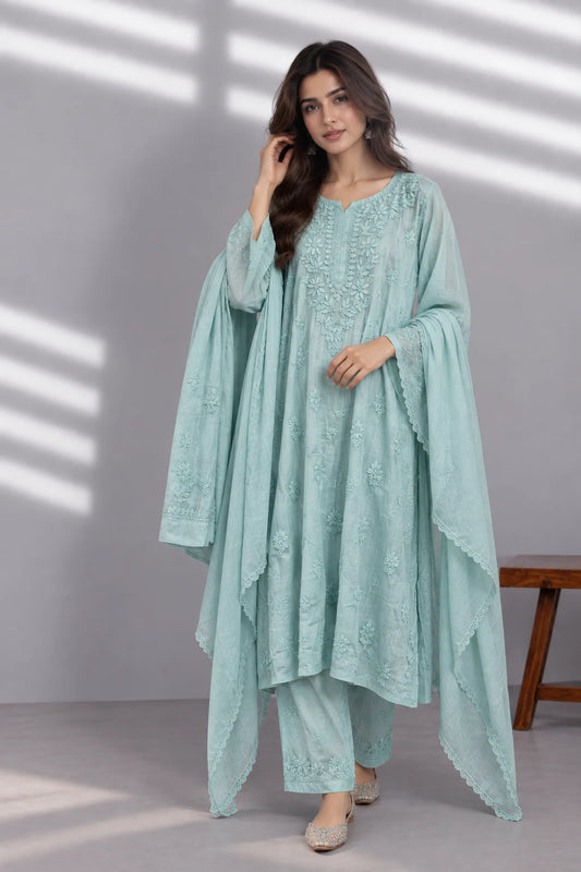 A woman models a light mint green embroidered traditional outfit with a matching draped outer layer, set against a gray wall with soft window shadow patterns.