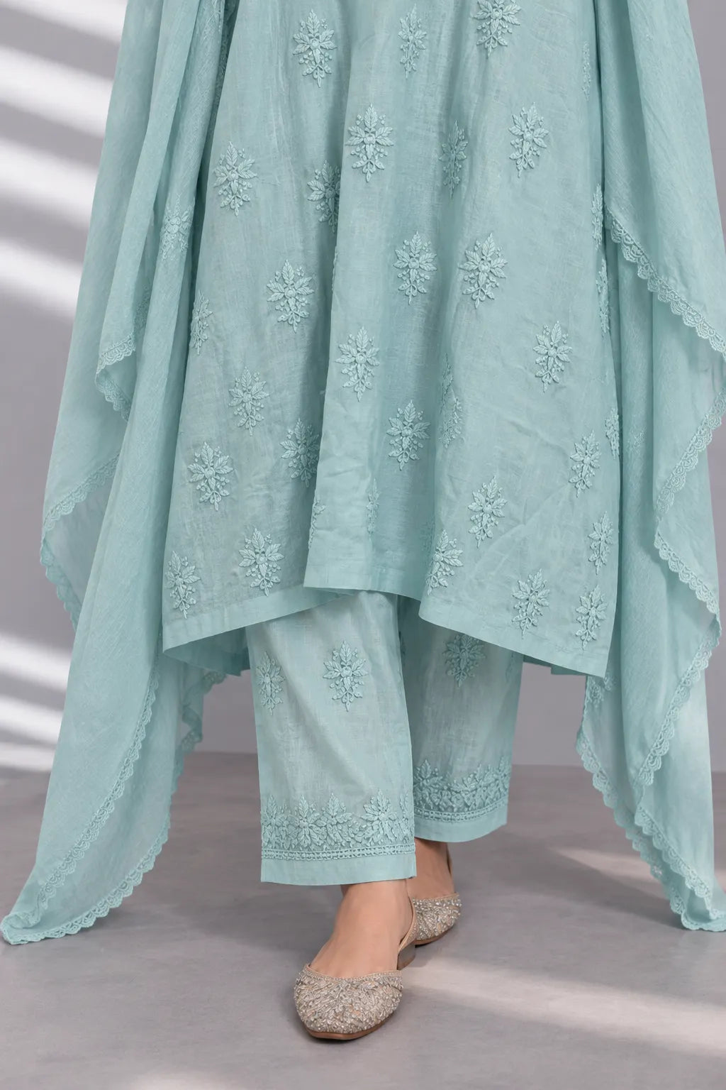 A close-up of a person wearing a light mint green traditional outfit with matching embroidered pants and a draped dupatta, paired with beige flat shoes.