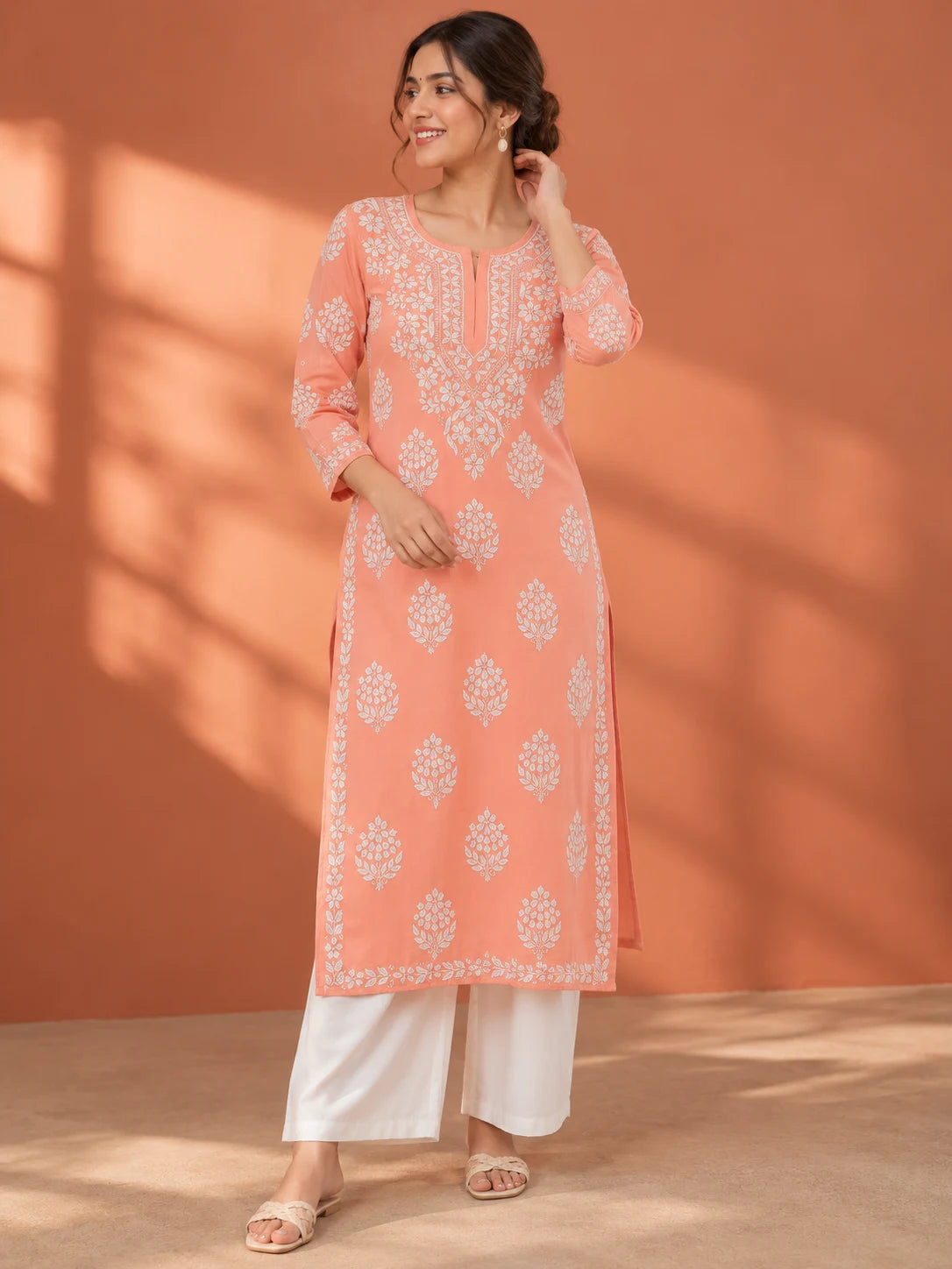 A woman models a peach-colored kurta with white floral embroidery, paired with white wide-leg pants, set against a warm terracotta background with soft window shadow patterns.