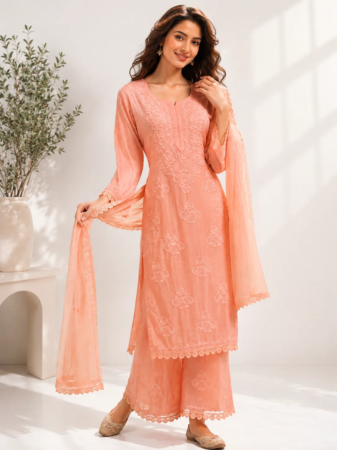 A woman models a peach-colored traditional Indian salwar kameez suit with matching dupatta. The outfit features intricate floral embroidery on the tunic and wide-leg pants, styled with beige flat shoes in a bright, modern room setting.