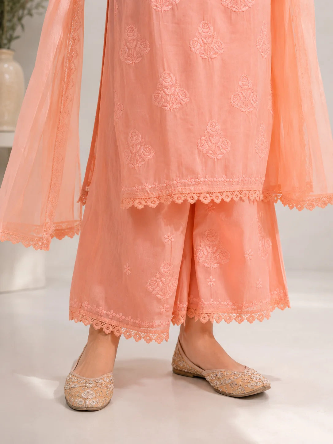 A close-up of a person wearing a peach-colored traditional outfit with embroidered details, lace-trimmed hems, sheer sleeves, and beige embellished flat shoes.