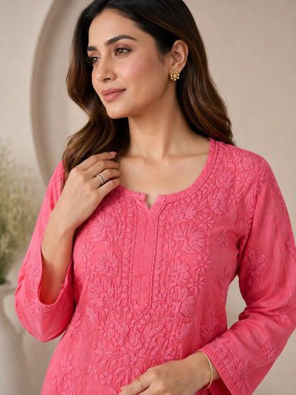 A woman wearing a bright pink embroidered kurta with a split neckline and long sleeves, shown in a softly lit indoor setting.
