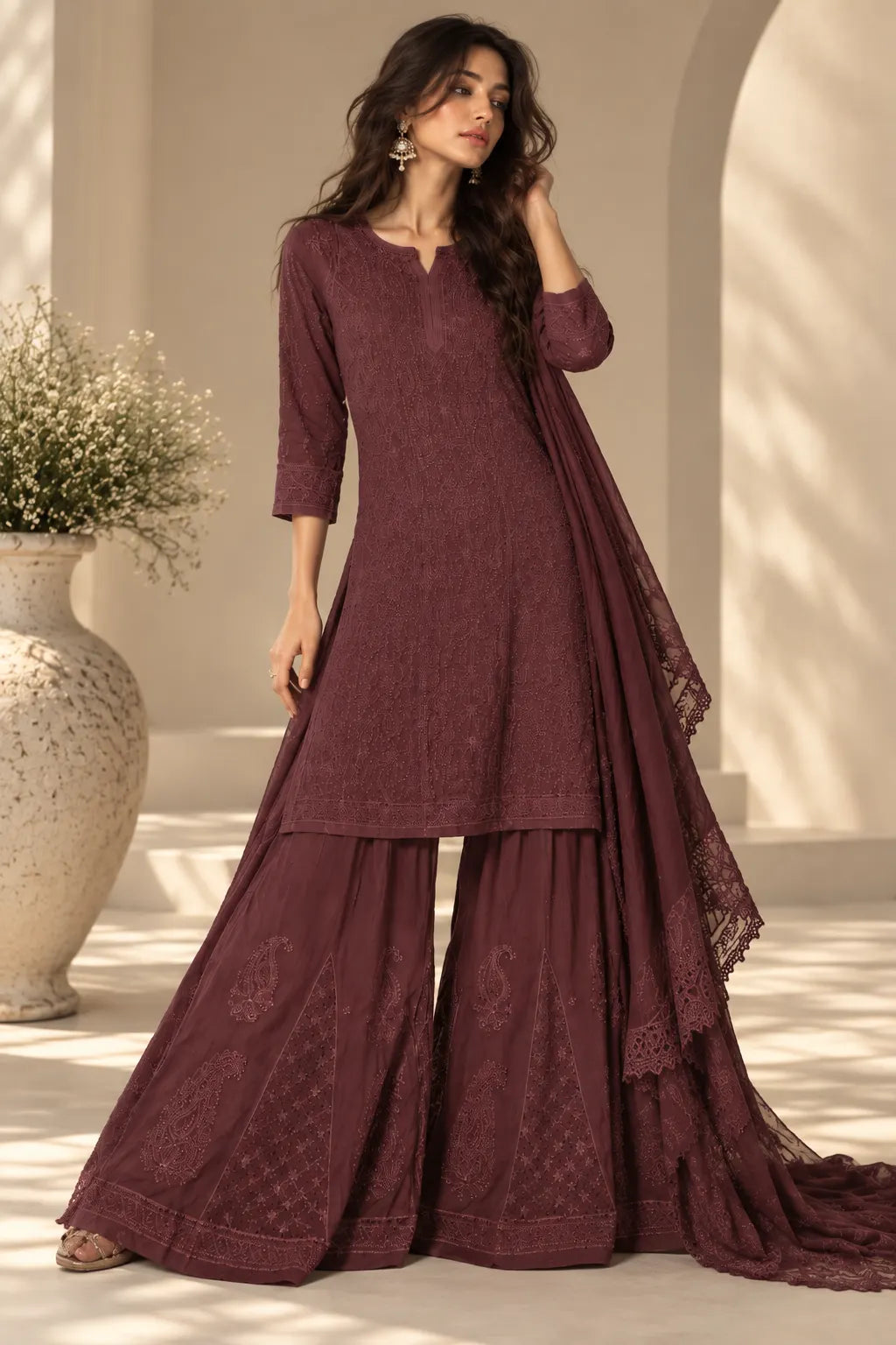 A woman models a three-piece maroon ethnic salwar kameez suit with matching dupatta in a sunlit architectural setting. The set features an embroidered kurta, wide flared pants, and a draped dupatta.