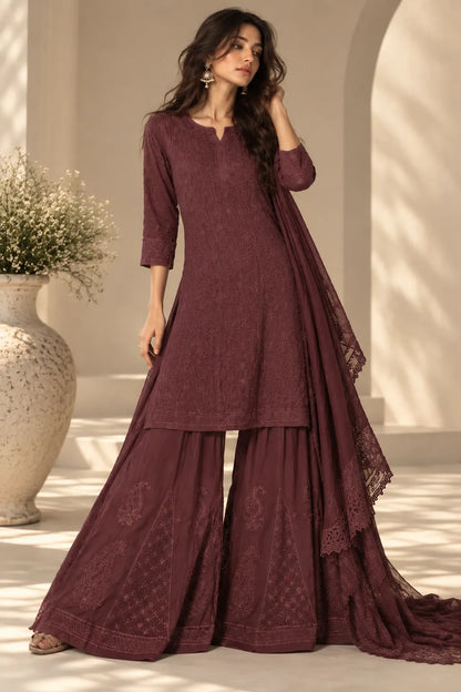 A woman models a three-piece maroon ethnic salwar kameez suit with matching dupatta in a sunlit architectural setting. The set features an embroidered kurta, wide flared pants, and a draped dupatta.