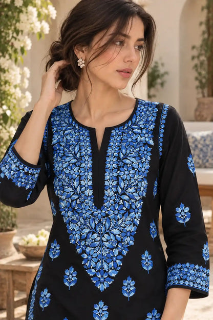 A woman wearing a black kurta with intricate blue floral embroidery on the front panel and cuffs, photographed in a softly lit outdoor setting with plants and furniture in the background.