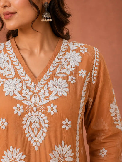 A close-up of a woman wearing a terracotta orange V-neck kurta with intricate white floral embroidery, paired with silver jhumka earrings, set against a matching terracotta background.