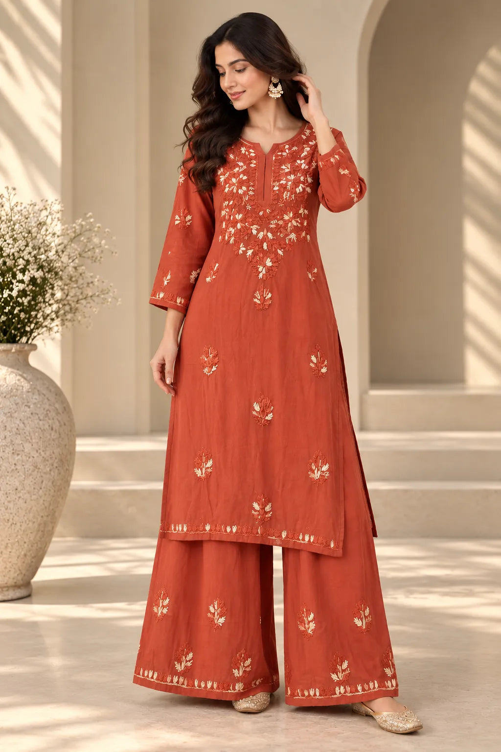 A woman models a rust-orange kurta and wide-leg palazzo pants set with white floral embroidery, set against a warm beige architectural background with a large vase of white flowers.