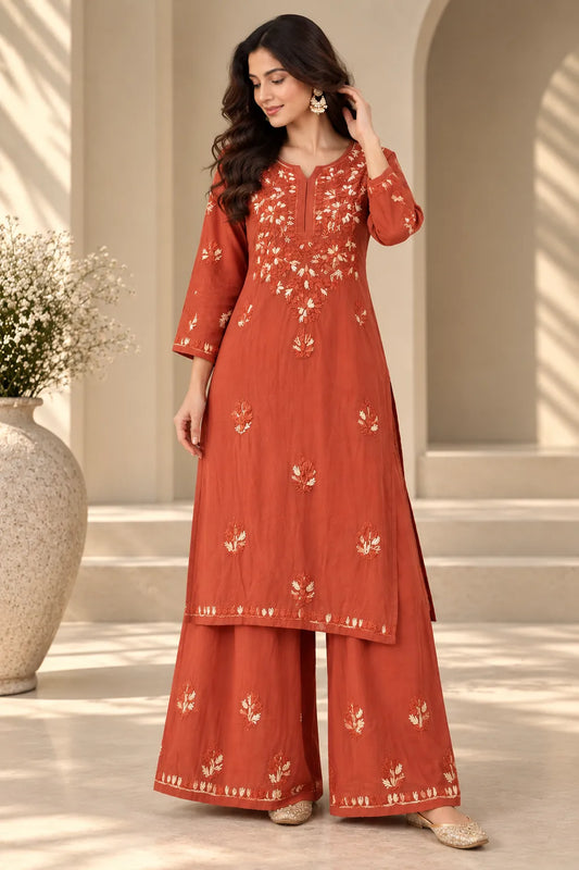 A woman models a rust-orange kurta and wide-leg palazzo pants set with white floral embroidery, set against a warm beige architectural background with a large vase of white flowers.