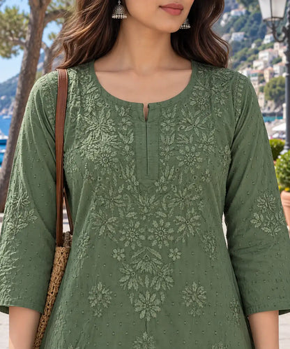 A woman wearing a sage green embroidered kurta with floral lace detailing, photographed outdoors near a coastal town with mountains and water in the background.