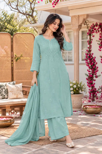 A woman models a mint green ethnic three-piece suit with a matching dupatta in an outdoor patio setting. The kurta features delicate floral embroidery, paired with matching wide-leg pants and a flowing dupatta.