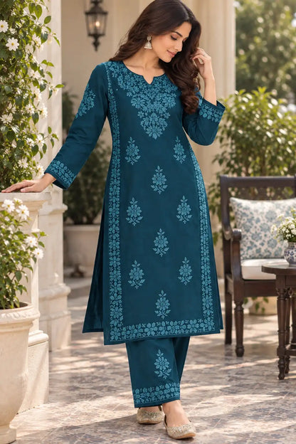 A woman models a teal blue ethnic salwar kameez suit with intricate light blue embroidery, standing on a stone patio with plants and a wooden chair in the background.