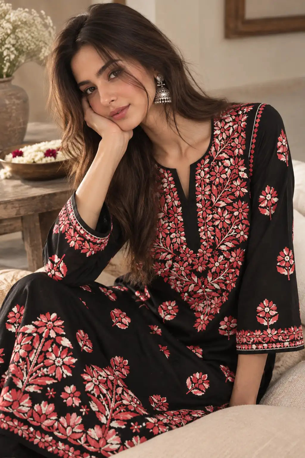 A woman wearing a black embroidered kurta with pink and red floral patterns, sitting on a beige sofa in a warm, softly lit indoor setting.