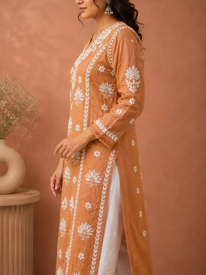A woman wearing a peach-orange embroidered kurta with white floral and geometric patterns, paired with a white inner dress, standing against a warm terracotta wall.
