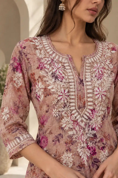 A close-up of a woman wearing a floral embroidered tunic top in dusty pink and purple tones with white embroidery details around the neckline and sleeves.