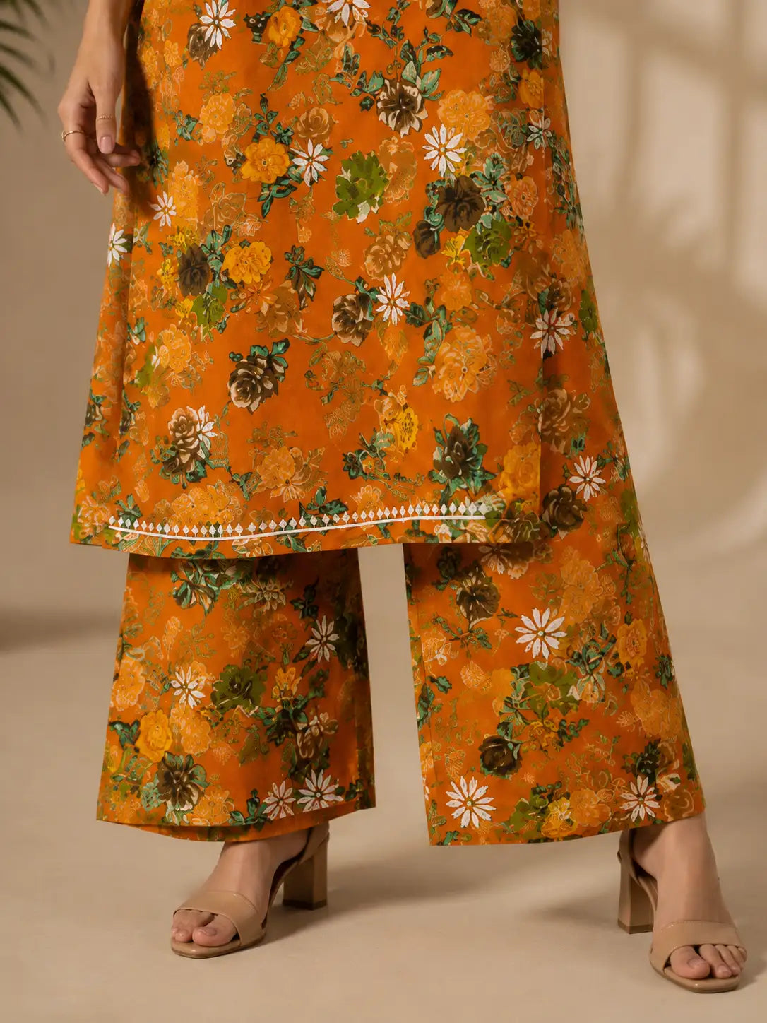 A person wearing a mustard-orange floral kurta and matching wide-leg pants with beige heeled sandals, shown from the waist down against a warm beige studio background.